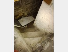 Eine enge Kellertreppe aus Beton, umgeben von unverputzten Ziegelwänden. Auf einer Stufe liegen ein umgestürztes weißes Brett und ein Paar Schuhe.