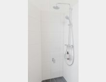 Ecke einer modernen Dusche mit weißen Wandfliesen, einem Glas-Duschabtrennung, einem verchromten Duschkopf und Handbrause. Ein kleiner Korb mit Pflegeprodukten ist an der Duschstange befestigt, der Boden besteht aus dunklen Fliesen.