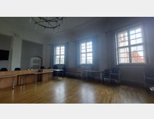 Ein heller Raum mit hohen Decken, drei großen Fenstern und einer blendenden Sicht nach draußen. Der Raum hat Holzböden, eine hölzerne Tischreihe entlang der Wand, mehrere Stühle und einen Kronleuchter an der Decke.