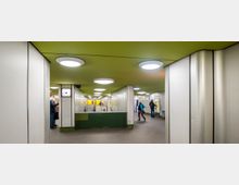 Unterirdische U-Bahnstation mit grüner Decke, rundlichen Deckenleuchten und grauen Wandverkleidungen. Im Vordergrund stehen Ticketautomaten, eine analoge Uhr ist an einer Säule angebracht, und mehrere Personen befinden sich im Raum.