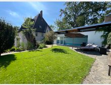 Haus mit Satteldach und Spitzgiebel, daneben ein moderner Anbau mit weißer Fassade, großen Fenstern und angrenzendem Innenpool. Im Vordergrund liegt ein gepflegter Rasen, umgeben von Bäumen, Pflanzen und einer gepflasterten Terrasse mit Sonnenliegen und einem Sonnenschirm.