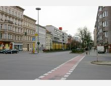 Straßenecke in einer Stadt mit mehrstöckigen Wohn- und Geschäftshäusern im Hintergrund, ergänzt durch einen Fahrradstreifen und Verkehrsampeln. Ein Schild weist auf eine U-Bahn-Station hin, während einige Fußgänger und parkende Autos sichtbar sind.