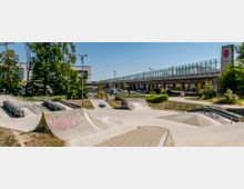 Moderner Skatepark mit verschiedenen Rampen, Rails und Betonelementen, umgeben von grünen Bäumen und Grasflächen. Im Hintergrund verläuft eine erhöhte Straßenbrücke mit Glasgeländer, daneben stehen vereinzelte Gebäude.