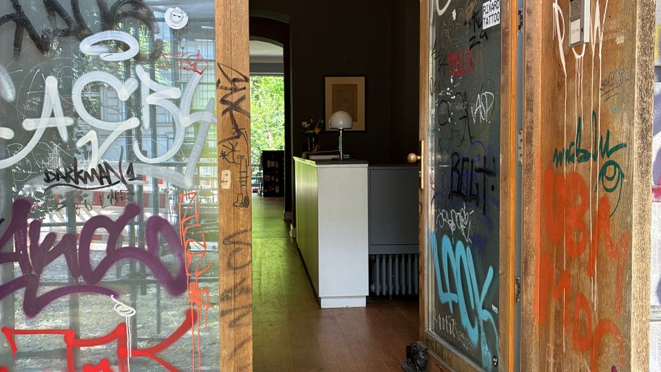 Eingangstür mit Holzrahmen und großen Glasflächen, die dicht mit Graffiti und Tags besprüht sind. Durch die offene Tür ist ein schmaler Innenraum mit Holzboden, dunklen Wänden und einem hellen Tresen sowie Lampen sichtbar.