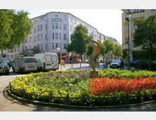 Ein runder Platz mit einer bepflanzten Blumenrabatte in der Mitte, die mit gelben, roten und violetten Blumen gestaltet ist, und einer goldfarbenen Skulptur. Im Hintergrund stehen mehrstöckige Wohngebäude, geparkte Autos und Gehwege mit Bäumen sowie ein kleines Café mit Außensitzplätzen.