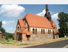 Backsteinkirche mit rotem Ziegeldach und einem hohen, spitzen Kirchturm, erbaut auf einem Sockel aus Naturstein. Die Kirche liegt an einer Straße, umgeben von Bäumen und einer ländlichen Umgebung.
