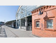 Bahnhofsgebäude mit einer Kombination aus Glas- und Ziegelarchitektur. Links ein Bahnsteig mit Überdachung und rechts eine Backsteinfassade mit vergitterten Fenstern und einer Wandlampe; im Hintergrund blaue Himmelsicht.