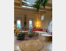 Gemütlicher Wohnraum mit einer großen, niedrig angesetzten, gepolsterten Sitzecke in Erdtönen und bunten Kissen. Der Raum hat gewölbte Fenster mit Steinverkleidung, hängende Dekoelemente, runde Teppiche aus Naturmaterialien und einen Glasdachbereich, der natürliches Licht hereinlässt.