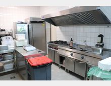 Moderne Großküche mit Edelstahlarbeitsflächen, Gaskochfeldern, einer großen Dunstabzugshaube und einem Kühlschrank. Im Raum befinden sich weiße Fliesenwände, Regale mit Geschirr und Behälter zur Lebensmittelaufbewahrung.