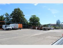 Eine große, offene Betriebsfläche mit mehreren abgestellten Fahrzeugen, darunter weiße Lieferwagen, ein orangefarbener Gabelstapler und ein Anhänger. Im Hintergrund Bäume, Metallregale und ein Gebäude am Rand des Grundstücks.