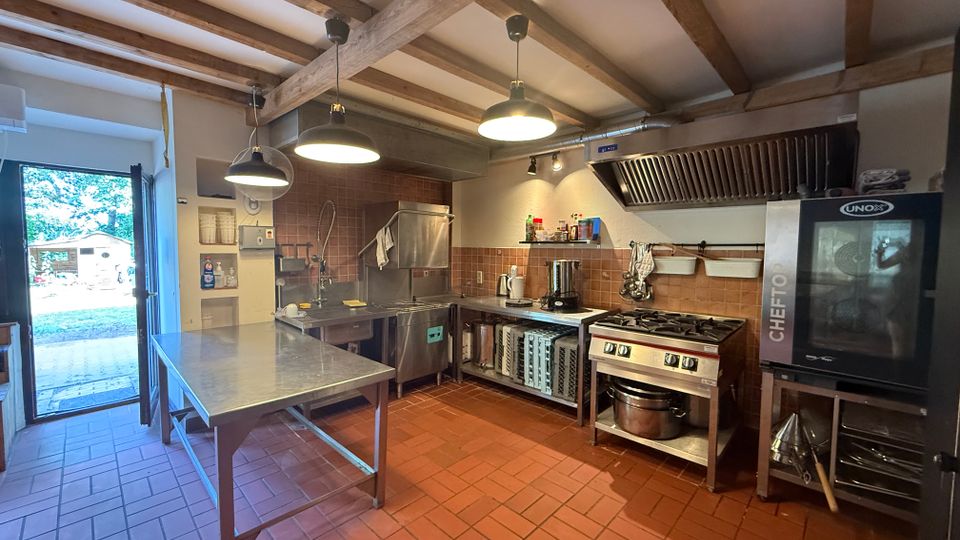 Großküche mit terrakottafarbenen Fliesen, sichtbaren Holzbalken an der Decke und hängenden Industrielampen. Zu sehen sind ein Edelstahl-Arbeitstisch, Spülbereich, Herd mit großer Dunstabzugshaube sowie ein hoher Kombidämpfer; links führt eine offene Tür nach draußen.