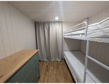 Kleines Schlafzimmer mit hellen Holzvertäfelungen an Wänden und Decke sowie Holzfußboden. Rechts steht ein weißes Metall-Etagenbett, links eine Kommode mit Holzplatte; am Ende des Raums hängt ein grauer Vorhang vor dem Fenster.