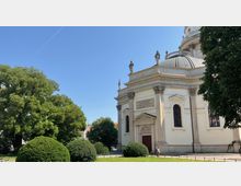 Historisches Kirchengebäude mit heller Fassade, einer Kuppel und dekorativen Statuen auf dem Dach. Im Vordergrund ein gepflegter Park mit Bäumen und Sträuchern unter klarem, blauem Himmel.
