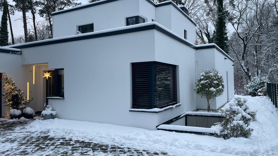 Modernes, weiß verputztes Flachdachhaus mit kubischer, gestufter Architektur und großen Fenstern mit dunklen Außenjalousien. Im verschneiten Vorgarten liegen ein gepflasterter Weg, niedrige Mauern und Sträucher; dahinter steht ein Wald aus hohen Bäumen.