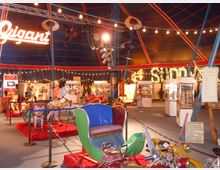 Innenraum eines Zirkuszelts mit einer Ausstellung von historischen Karussellfahrzeugen, Schautafeln und dekorativen Lichtern. Im Hintergrund sind beleuchtete Schriftzüge und Ausstellungsstücke sichtbar, während der Raum von Leuchtketten und einem blauen Zeltdach mit roten Streifen überspannt wird.