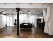 Industrieller Innenraum mit rustikalem Betonboden, einer schwarzen Couchgarnitur vor weißen Vorhängen und einem schwarzen Metallpfeiler in der Mitte. Rechts befindet sich eine Bar mit schwarzen Barhockern, links ein Schreibtisch mit Spiegel.