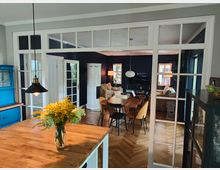 Offener Wohnbereich mit einem Übergang vom Esszimmer ins Wohnzimmer, unterteilt durch eine Glastrennwand mit weißen Rahmen. Im Vordergrund ein Holztisch mit einem Blumenstrauß, im Hintergrund ein Holzfußboden in Fischgrätoptik, ein runder Kachelofen und gemütliche Sitzmöbel.