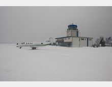 Flughafen bei winterlicher Schneelandschaft mit einem kleinen weißen Flugzeug auf dem Rollfeld und einem Kontrollturm im Hintergrund. Der Himmel ist grau, und das Gelände ist mit Schnee bedeckt.