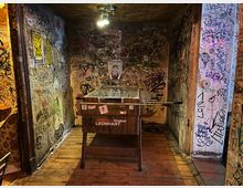 Schmaler Innenraum wie ein Flur in einer Bar oder einem Klub, mit Holzboden und einer Deckenlampe über einem zentral stehenden Kickertisch. Die Wände und Türrahmen sind dicht mit Graffiti, Stickern und Postern bedeckt.