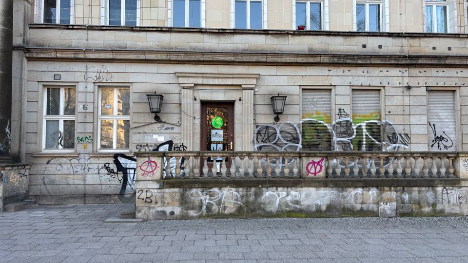 Straßenfassade eines mehrgeschossigen Steinbaus mit zentraler Eingangstür, zwei Wandlaternen und einer niedrigen Balustrade vor dem Gebäude. Mehrere Fenster mit Rollläden sowie großflächige Graffiti und Schmierereien sind an der Wand und am Sockel zu sehen.