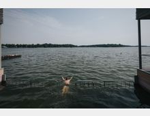 Ein Mann schwimmt in einem See, der von einem bewaldeten Ufer umgeben ist. Im Vordergrund befinden sich Metallgeländer am Wasser, und links ist ein Steg mit Sonnenbadenden zu sehen.