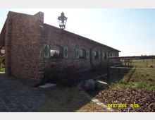 Backsteinbau mit niedrigen Fenstern, die mit grünen Fensterläden verziert sind, und einem an der Wand befestigten Laternenlicht. Im Vordergrund führt ein gepflasterter Weg entlang blühender Rosen, während im Hintergrund eine offene Wiesenlandschaft mit Holzpfosten und gespannter Schnur sichtbar ist.