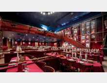 Großer Veranstaltungsraum mit rotem Teppich und gedeckten Tischen, eingerahmt von einer Galerie mit roten Wänden, dekorativen Theaterplakaten und einer geschwungenen Treppe. Die Decke ist dunkelblau mit Sternenlichtern gestaltet, und die Bühne ist im Hintergrund erkennbar.