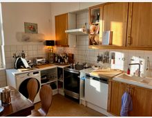 Kleine Küche mit Holzmöbeln und weißen Fliesen, ausgestattet mit Herd, Spülmaschine und Spüle. Auf der Arbeitsplatte stehen Küchenutensilien, Messer sind an einer Magnetleiste befestigt, und ein Tisch mit Stühlen befindet sich im Vordergrund.