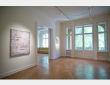 Heller Raum mit Parkettboden, großen Fenstern und weiß gestrichenen Wänden, an denen abstrakte Kunstwerke hängen; im Hintergrund ein weiterer Raum mit einem gelben Sofa und reich verzierten Fenstern.