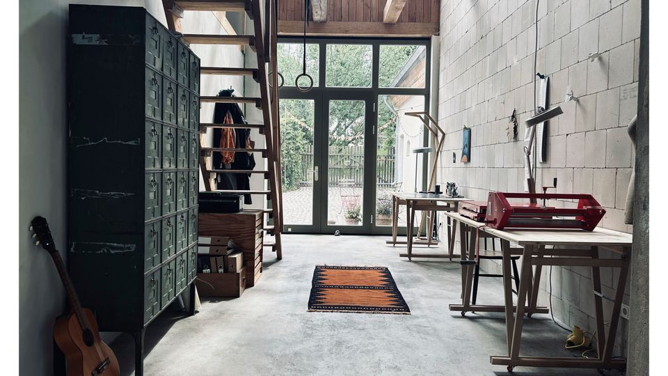 Großer Werkstatt- oder Atelierraum mit Betonboden, Holztreppe und großen Glastüren, die auf einen begrünten Außenbereich führen. Im Raum stehen Arbeitsbänke und Tische, daneben ein großes dunkelgrünes Metallschrank-Element, ein kleiner Teppich und eine an die Wand gelehnte Gitarre.