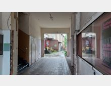 Ein überdachter Durchgang mit gefliesten Boden führt zu einem Hinterhof. Im Hintergrund sichtbar sind ein rotes Backsteingebäude, eine kleine Treppe mit Topfpflanzen und eine bepflanzte Umgebung, umgeben von Gebäuden.