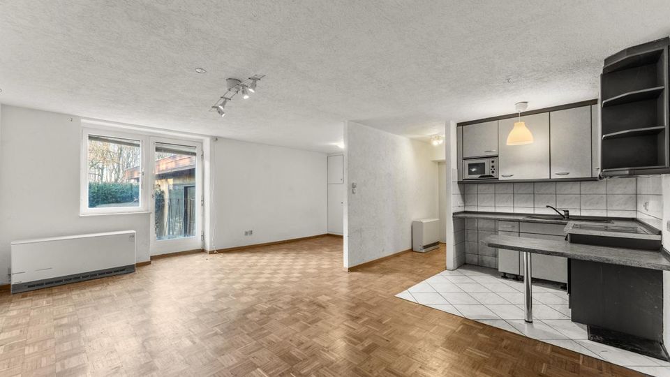 Heller, leerer Wohnraum mit Parkettboden und weißen Wänden, mit großem Fenster und Glastür zum Balkon. Rechts befindet sich eine offene Küchenzeile mit grauen Schränken, gefliester Rückwand und kleinem Tresen auf einem gefliesten Bodenbereich.
