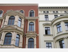 Fassadenansicht historischer Wohngebäude mit dekorativen, hell verputzten Erkern und Säulen auf rotem Ziegel- und weißem Putzuntergrund. Große Rundbogenfenster und florale Stuckelemente prägen die architektonischen Details.