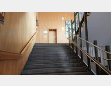 Eine Treppe in einem Innenraum mit Holzverkleidung an den Wänden und einem Geländer aus Metall. Am oberen Ende der Treppe befindet sich eine Tür und eine Wanduhr, daneben hängen Fotos und ein Schild.