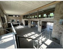 Großer, offener Innenraum in einem sanierten Industriegebäude mit unverputzten Ziegelwänden, hohen Decken und großen Fenstern. Eine Galerie verläuft entlang der oberen Etage, und Tageslicht fällt durch die Fenster auf den betonierten Boden.