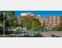 Industriegebäude mit roter Backsteinfassade und großem „VERITAS“-Schriftzug auf dem Dach, umgeben von Bäumen und einem grünen Zaun; im Hintergrund ein Turm mit großer Uhr. Der Vordergrund zeigt eine gepflasterte Fläche und einzelne Laternen.