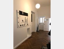 Flur mit weiß gestrichenen Wänden, einem dunklen Fischgrät-Holzboden und einer runden Deckenleuchte. An der Wand hängt ein abstraktes Gemälde in Schwarz-Weiß, und im Hintergrund sind weiße Türen und ein Staubsauger sichtbar.