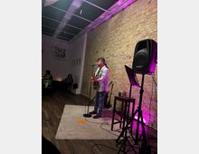 Small indoor performance space with an exposed brick wall, a rug-covered stage area, and purple accent lighting. A microphone stand, speaker on a tripod, music stand, and a stool with equipment sit beside the stage, with seating and a “Price & Son” sign visible at the back.