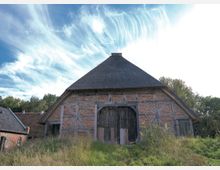 Historisches Fachwerkgebäude mit Backstein- und Holzelementen und einem Reetdach, umgeben von Wiesen und Bäumen; großes hölzernes Tor in der Mitte der Fassade.
