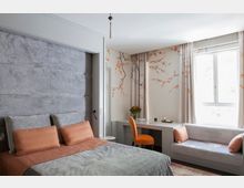 Ein modern eingerichtetes Schlafzimmer mit einem grauen gepolsterten Bettrahmen und orangen Akzentkissen. Die Wände sind mit einer Tapete mit Kirschblütenmotiv dekoriert, daneben ein Schreibtisch mit einem orangefarbenen Stuhl und ein gepolstertes Sofa vor einem großen Fenster.