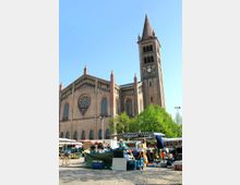 Backsteinkirche mit hohem Glockenturm und Rosettenfenster im Hintergrund, davor ein Marktplatz mit Ständen, die Lebensmittel und Waren anbieten, sowie Personen, die einkaufen. Im Vordergrund Kopfsteinpflaster und grüne Bäume an den Seiten.