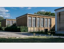 Flacher Gebäudekomplex mit gelben Putzfassaden und mehreren großen Glasbausteinfenstern, umgeben von Sträuchern und einem Metallzaun, im Hintergrund Bäume und blauer Himmel.