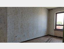 Ein leerer Raum mit vergilbter Tapete in floralem Muster, abgenutztem Holzboden und einem rechteckigen Fenster mit braunem Rahmen, das nach draußen auf Bäume und Gebäude blickt. Im Raum liegen ein paar Metallstreben sowie Schmutz auf dem Boden.