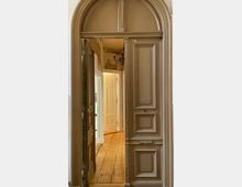 Eine geöffnete braune Holztür mit dekorativen Paneelen und rundem Oberlicht führt in einen Korridor mit Holzboden. Im Hintergrund sind weitere Türen und ein Kronleuchter sichtbar, die eine historische Innenarchitektur andeuten.