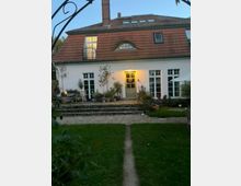 Zweistöckiges Wohnhaus mit weißer Fassade und einem roten Ziegeldach, das Dach hat Gaubenfenster und Dachfenster. Vor dem Haus befindet sich ein gepflegter Garten mit Rasen, einem gepflasterten Weg und mehreren Topfpflanzen auf einer erhöhten Terrasse.