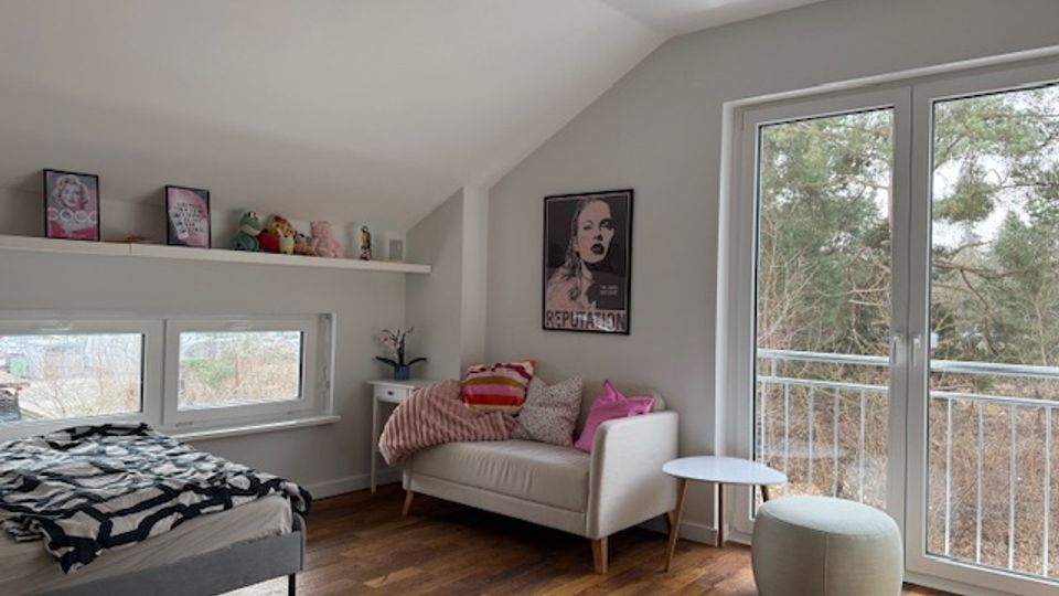 Bright attic bedroom with a sloped ceiling, wooden floors, a low bed beneath small horizontal windows and a long wall shelf. A white sofa with pillows sits by a large glass door to a balcony overlooking trees, with a small side table and round ottoman nearby.