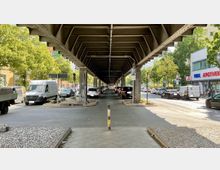 Straßenszene unter einer erhöhten Eisenbahnbrücke mit sichtbarer Stahlkonstruktion. Entlang der Straße parken Autos, flankiert von Gehwegen, Bäumen und Gebäuden wie einer Apotheke auf der rechten Seite.