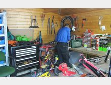 Eine Werkstatt mit Holzverkleidung, in der Werkzeuge an der Wand hängen und verschiedene Fahrräder sowie Reparaturmaterialien auf dem Boden und den Tischen verteilt sind. Eine Person steht an einer Werkbank und arbeitet an einem Objekt, umgeben von Werkzeugkästen und Regalen mit Zubehör.