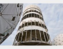 Blick auf eine verfallene Radaranlage mit runden, kuppelförmigen Strukturen und einem zylinderförmigen Turm mit zerrissenen Verkleidungen. Die weißen Kuppeln bestehen aus geometrischen Paneelen, einige mit Graffiti, während der Turm offene Fensterrahmen und sichtbaren Beton zeigt.