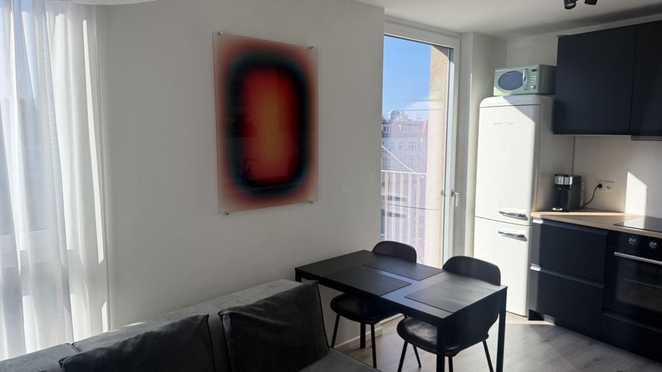 Heller Wohn- und Essbereich mit kleiner Küchenzeile, schwarzem Esstisch und Stühlen sowie dunklen Küchenschränken neben einem weißen Retro-Kühlschrank. Große Fensterfront mit Vorhängen und Glastür zum Balkon lassen viel Tageslicht herein; an der Wand hängt ein Bild.